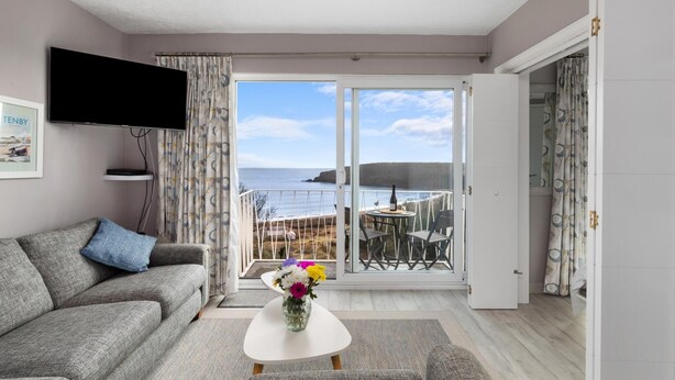 Uma sala de estar moderna com um sofá cinzento, uma televisão de ecrã plano, uma pequena mesa com um vaso de flores e uma varanda com vista para o mar.