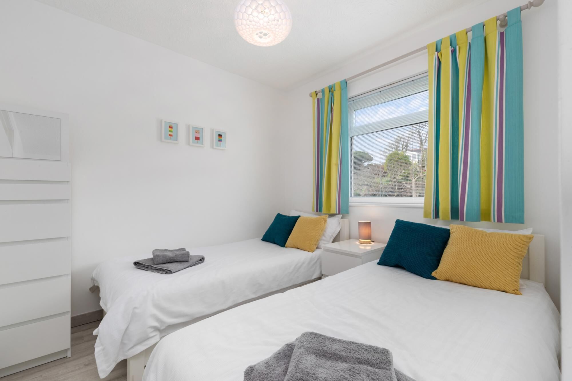 A bedroom with two beds, a window with striped curtains, a bedside table with a lamp, and a wardrobe.