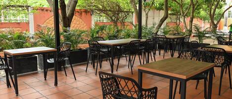 Un café al aire libre con varias mesas y sillas, rodeado de vegetación y un piso de baldosas.
