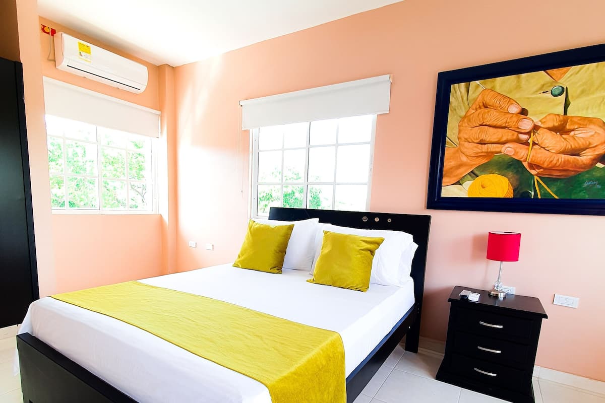 Un lit bien fait, recouvert d’une couverture jaune, avec deux oreillers, une table de chevet sur laquelle se trouve une lampe, et un tableau accroché au mur.