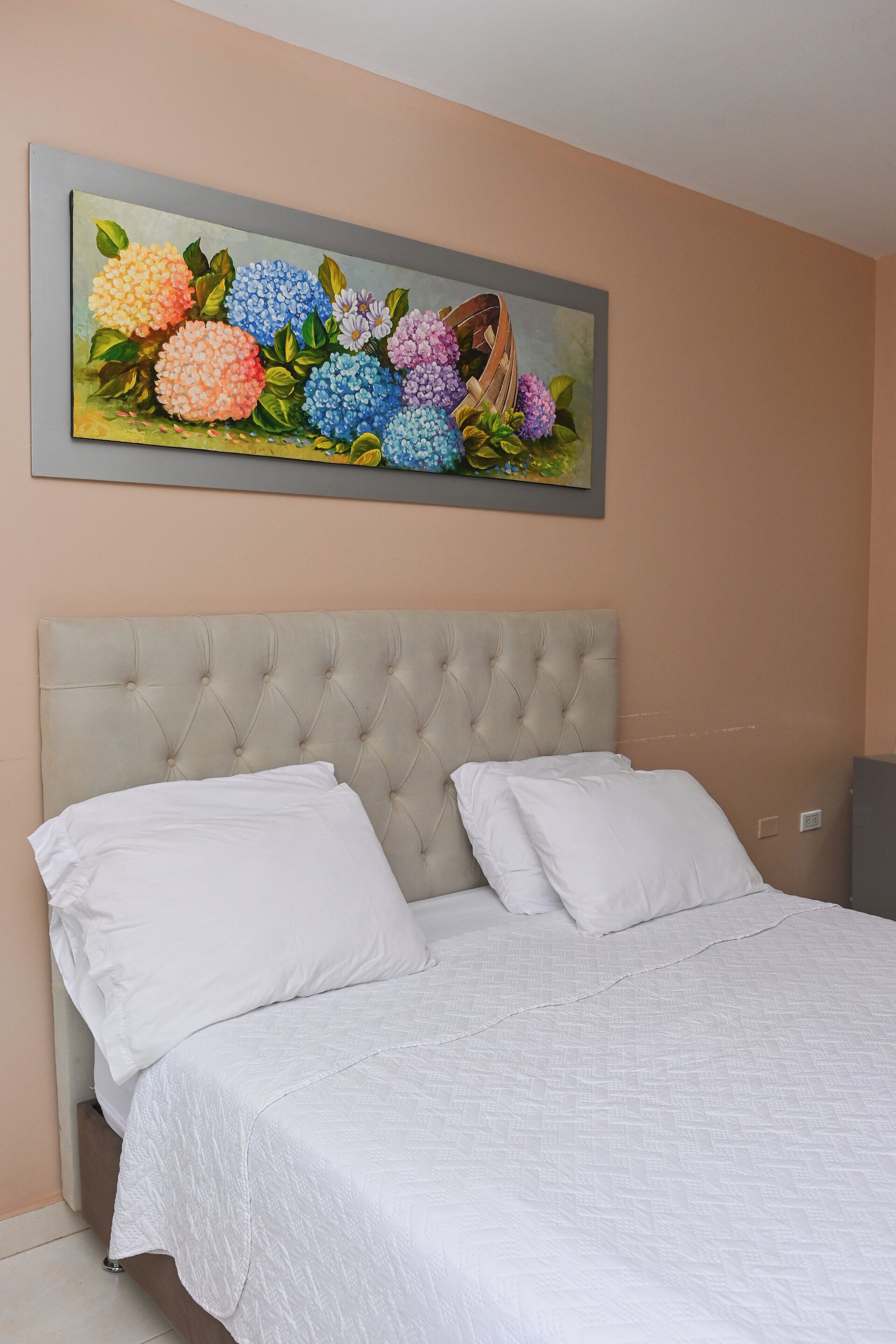A bedroom with a tufted headboard, white bedding, and a floral painting above the bed.