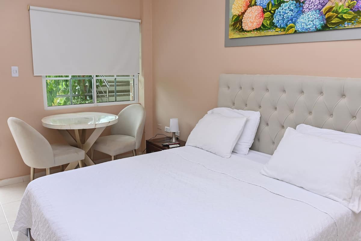 Une chambre avec un grand lit, une table ronde, deux chaises, une fenêtre donnant sur un paysage verdoyant et un tableau accroché au mur.