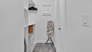 Un pasillo estrecho con una puerta blanca, una prenda negra colgada, una ventana con persianas y una tabla de planchar con estampado floral.