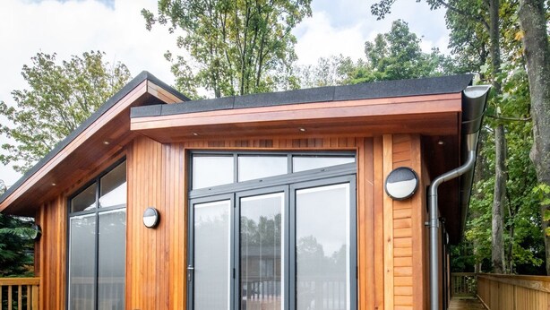 Une cabane moderne en bois, dotée de grandes portes-fenêtres coulissantes, d’une terrasse et d’un toit à forte pente.