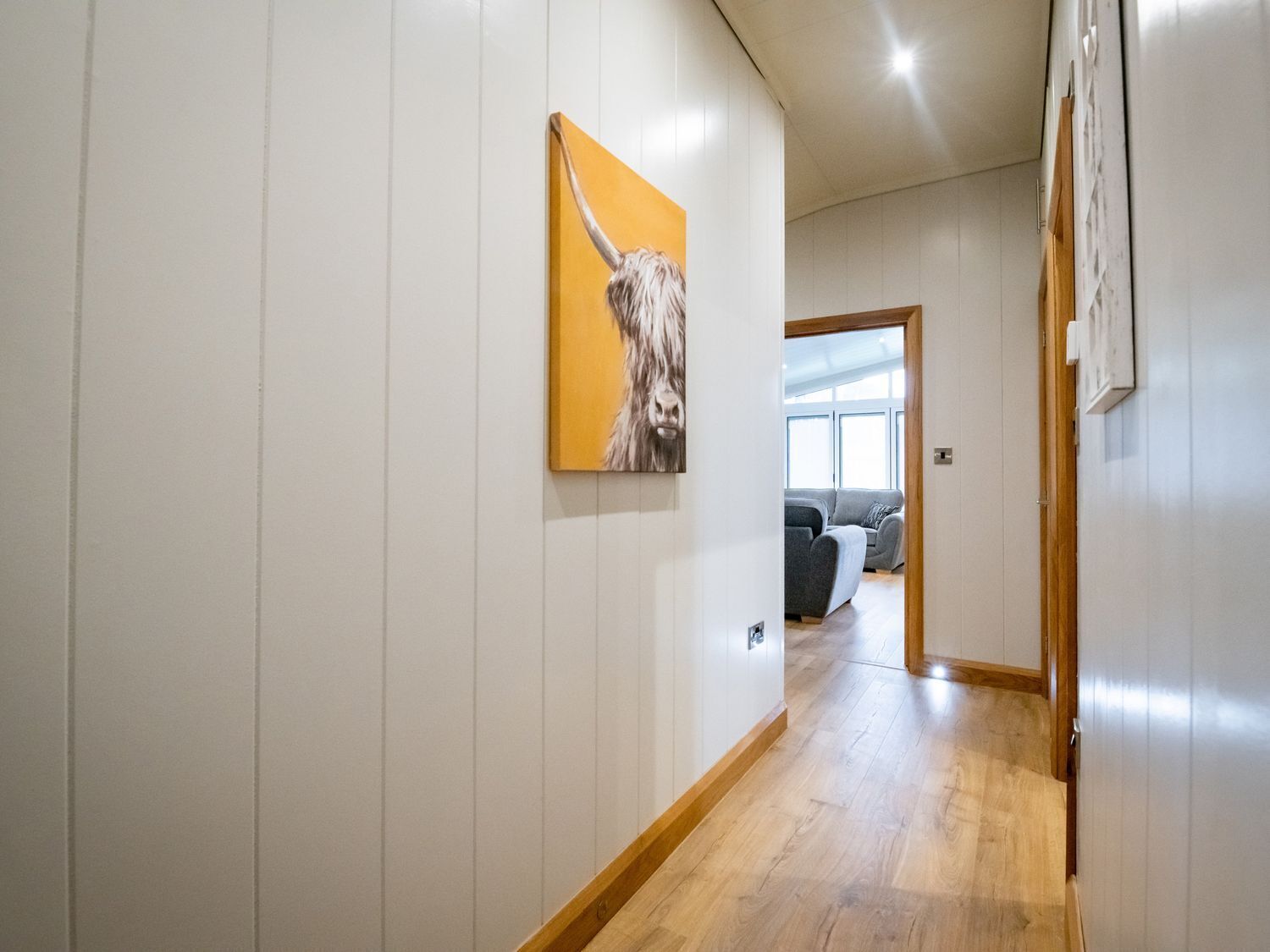 A hallway with white paneling and a wooden floor, leading to a room with a gray sofa and a window.