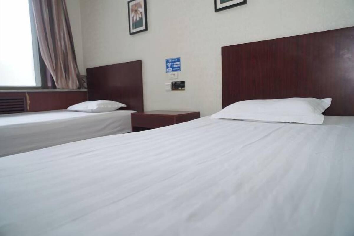 Ein Hotelzimmer mit zwei Einzelbetten, einem Holzkopfende, einem Nachttisch und einem Fenster mit Vorhängen.
