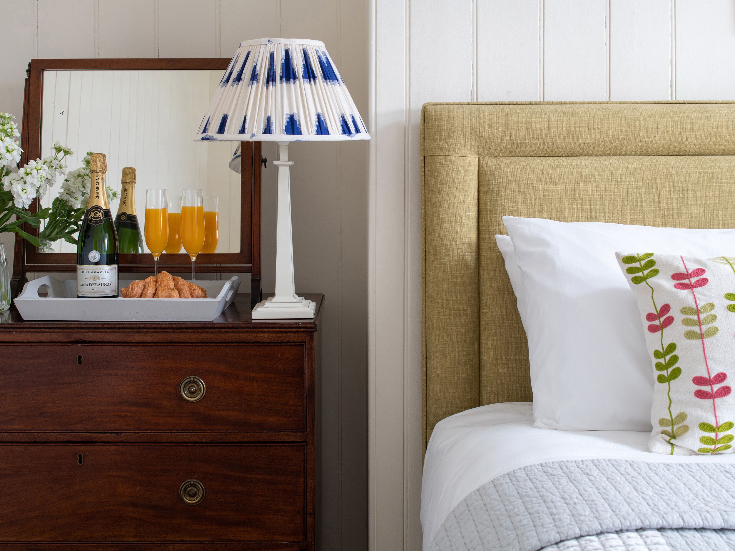 Habitación de hotel con cómoda de madera, lámpara, cama con cabecera beige y almohadón decorativo.