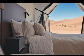 Una cama con ropa blanca y vistas a dunas del desierto a través de una ventana.