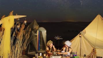 Un terrain de camping aménagé avec des tentes, une table dressée pour les repas, entouré d’éléments naturels et surplombé d’un ciel étoilé.