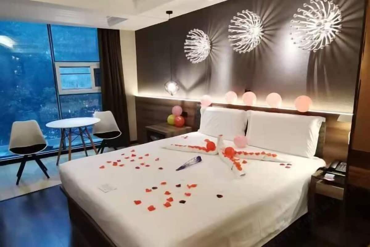 Ein Hotelzimmer mit einem großen Bett, einem kleinen Tisch, zwei Stühlen, einem Nachttisch und Blick auf die Stadt durch große Fenster.