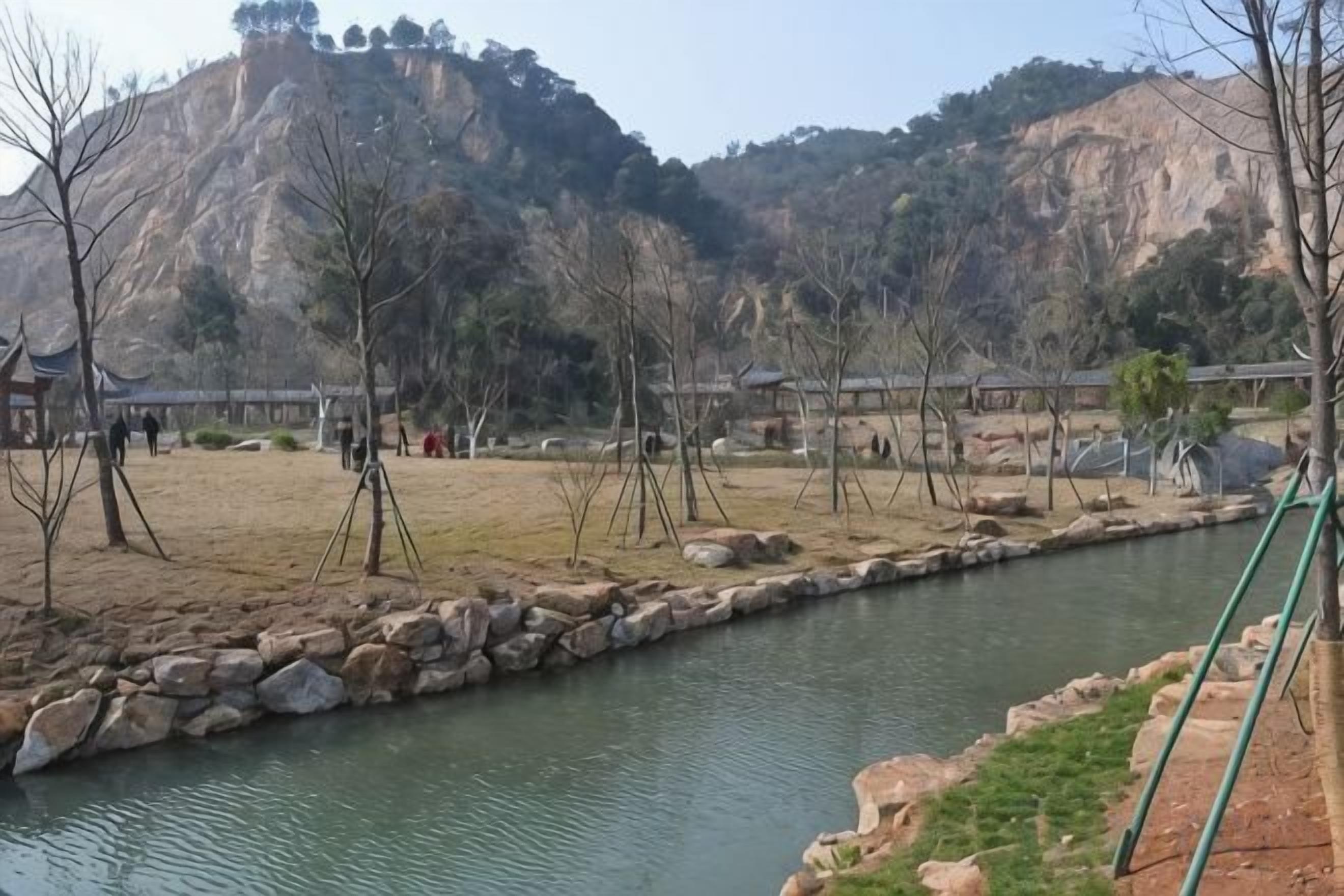 A riverside area with a stone pathway, a small bridge, and a hill in the background.