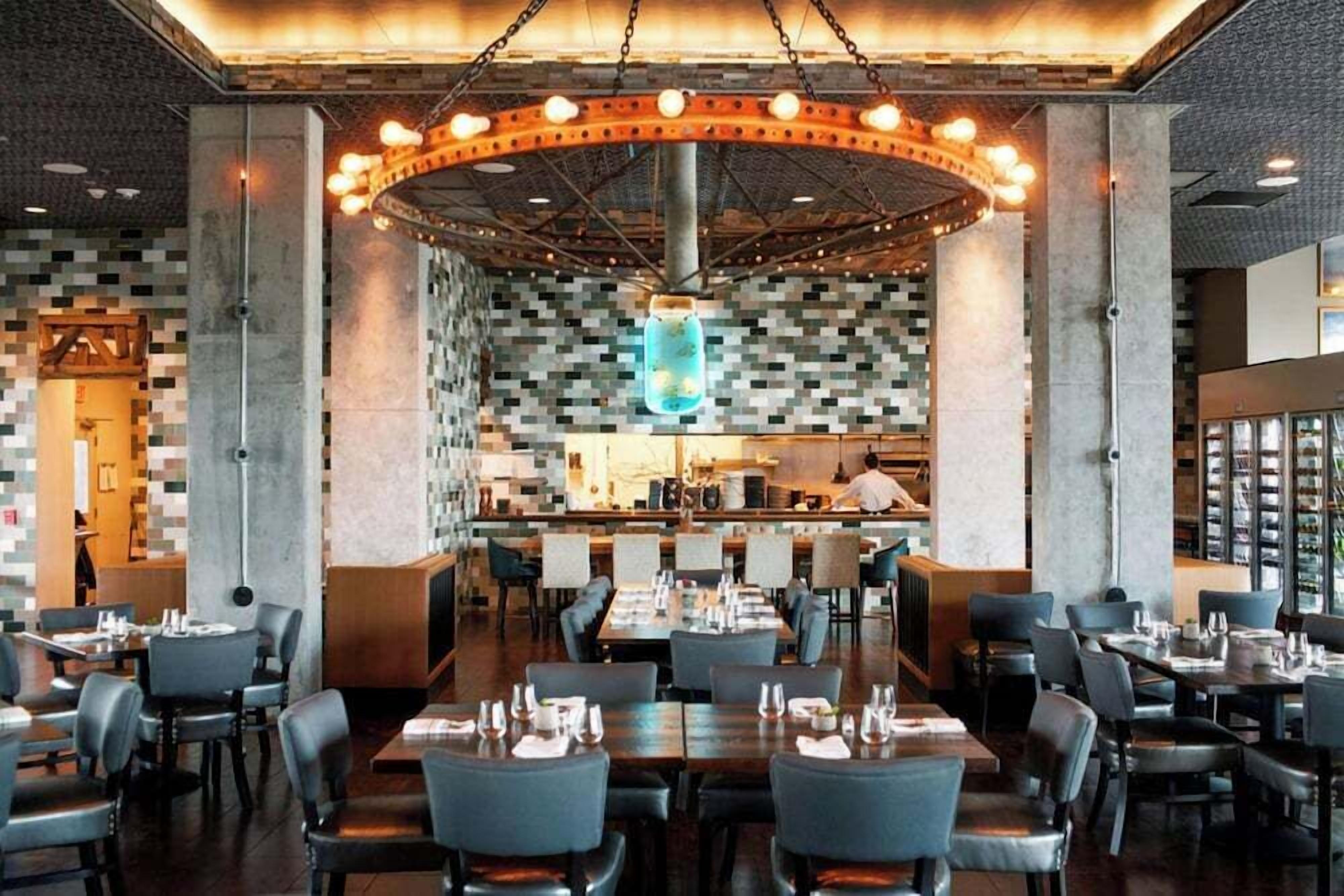 A modern restaurant with a central bar area, multiple dining tables, and a unique ceiling design.