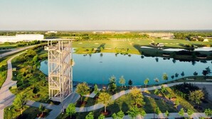 Un parcours de golf avec un grand étang, une haute structure métallique et un sentier menant à un bâtiment.