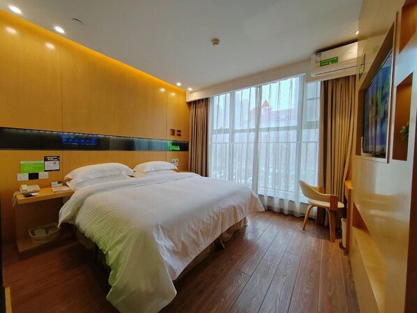 A hotel room with a large bed, a desk with a chair, a window with curtains, and an air conditioning unit.