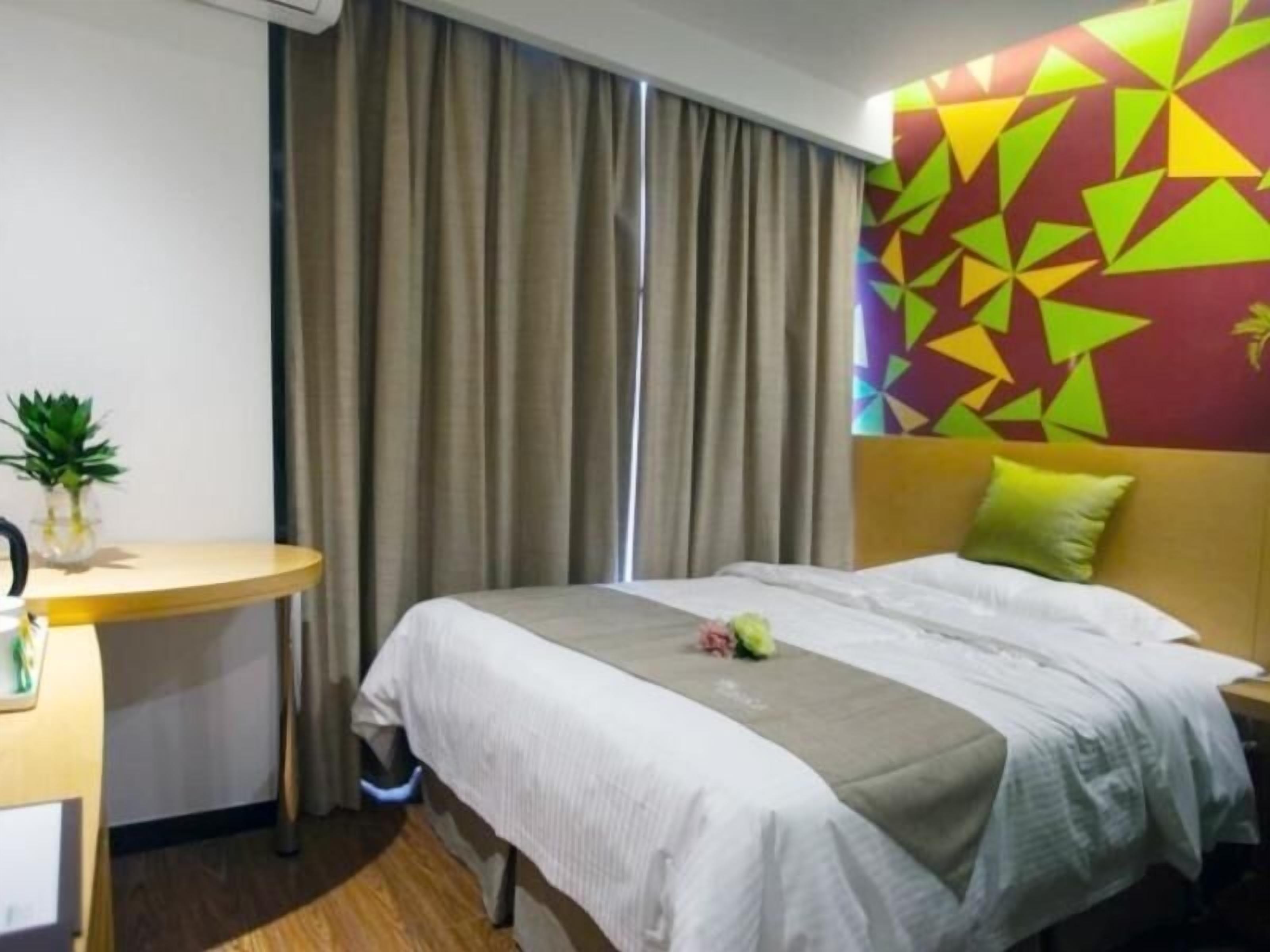 A hotel room with a bed, a desk, a potted plant, and a colorful geometric wall design.