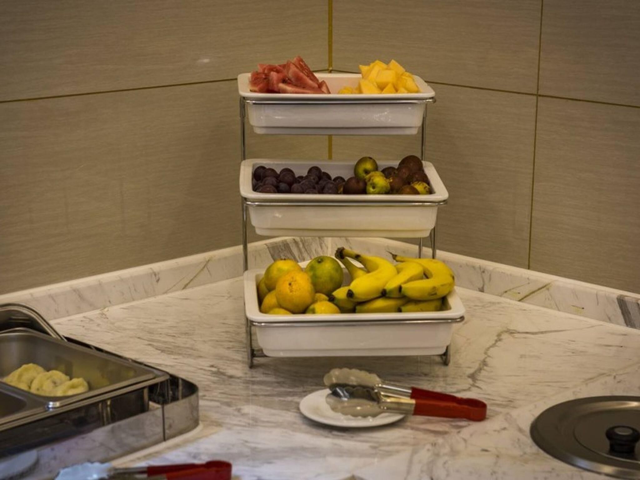 A buffet setup with trays of fruits and a fruit slicer on a marble countertop.
