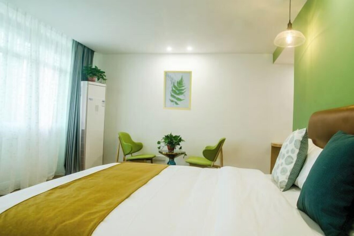 Hotellihuone, jossa on sänky, kaksi tuolia, pieni pöytä, jossa on kasvi, ja seinällä kehystetty taideteos.