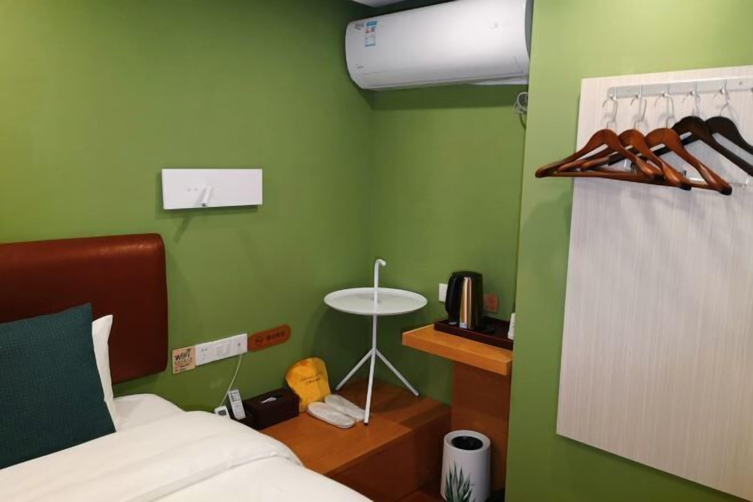 Habitación de hotel pequeña con cama, una mesita, un calentador de pared, un perchero y un cubo de basura.