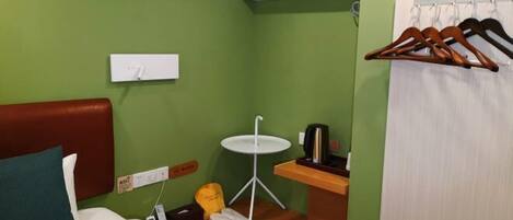 Habitación de hotel pequeña con cama, una mesita, un calentador de pared, un perchero y un cubo de basura.