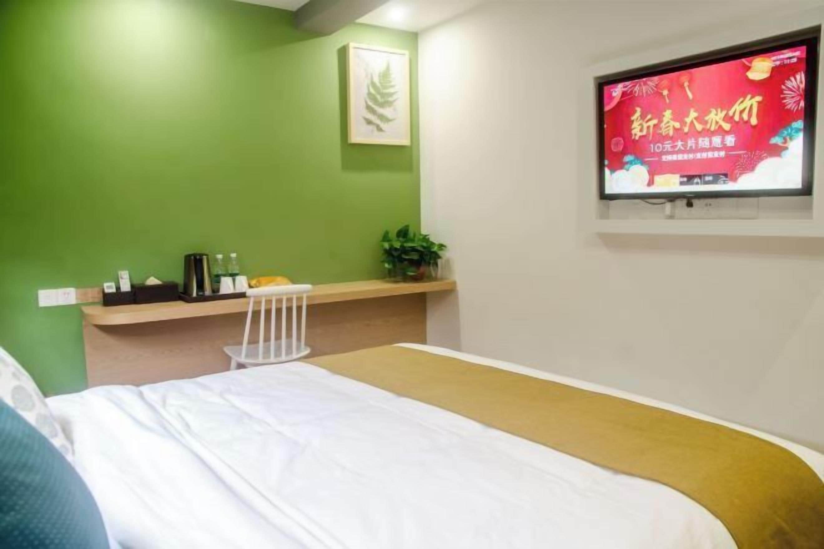 A hotel room with a bed, a small desk with a chair, a TV on the wall, and a green wall.