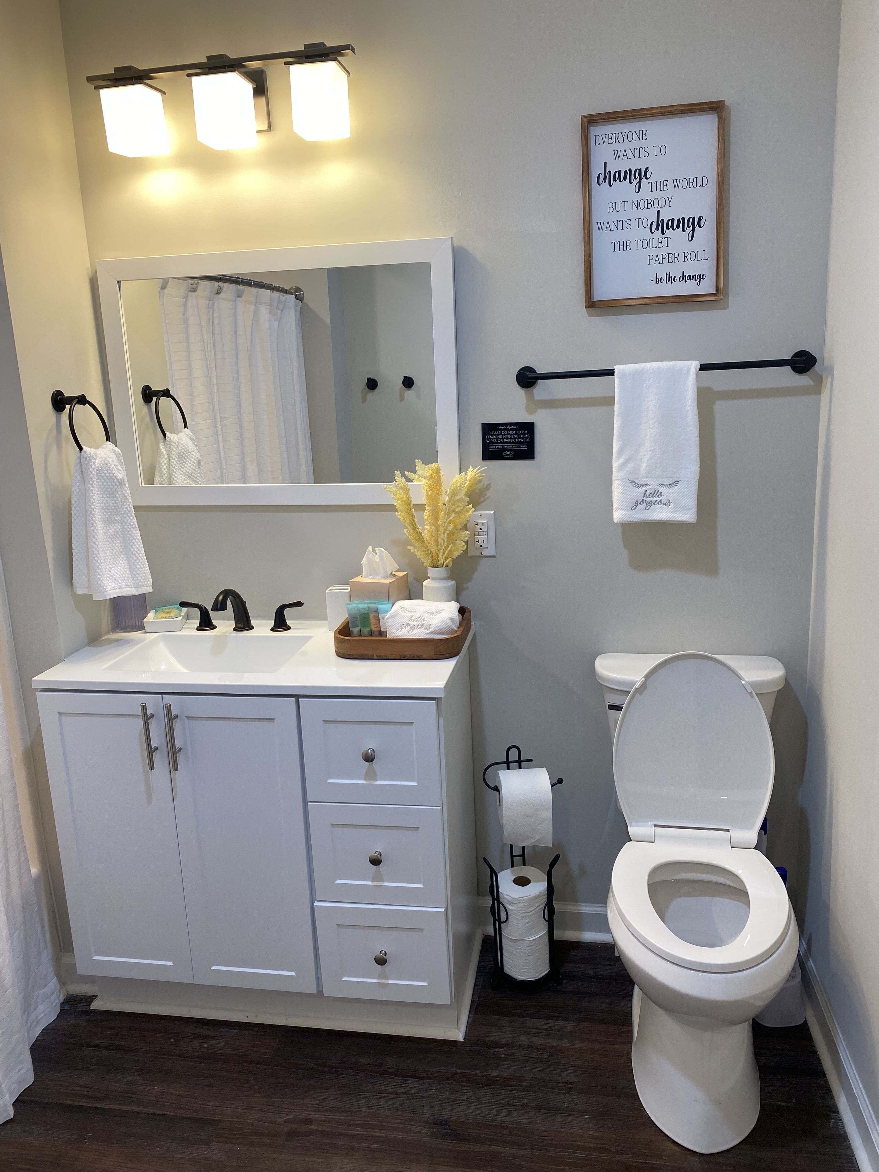 A bathroom with a white sink, mirror, and toilet. There are towels hanging on a rack, a framed quote on the wall, and a small plant on the sink.