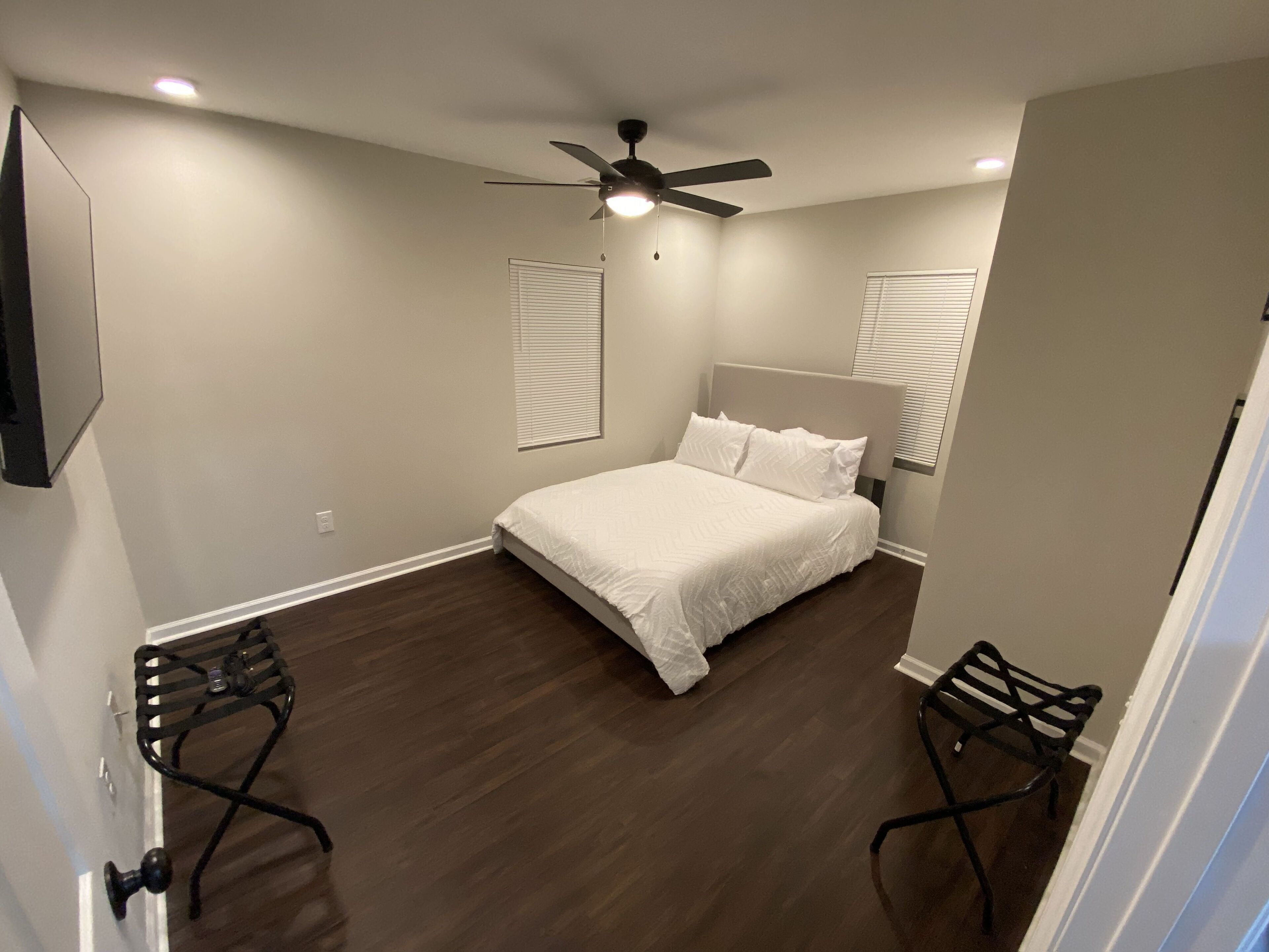 A bedroom with a bed, two chairs, a ceiling fan, and a TV mounted on the wall.