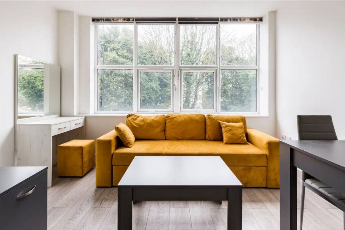 Un salon moderne avec un canapé jaune moutarde, une table basse noire et une grande fenêtre donnant sur la verdure.