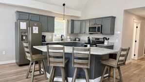 A modern kitchen with grey cabinets, a central island with bar stools, stainless steel appliances, and a pendant light.