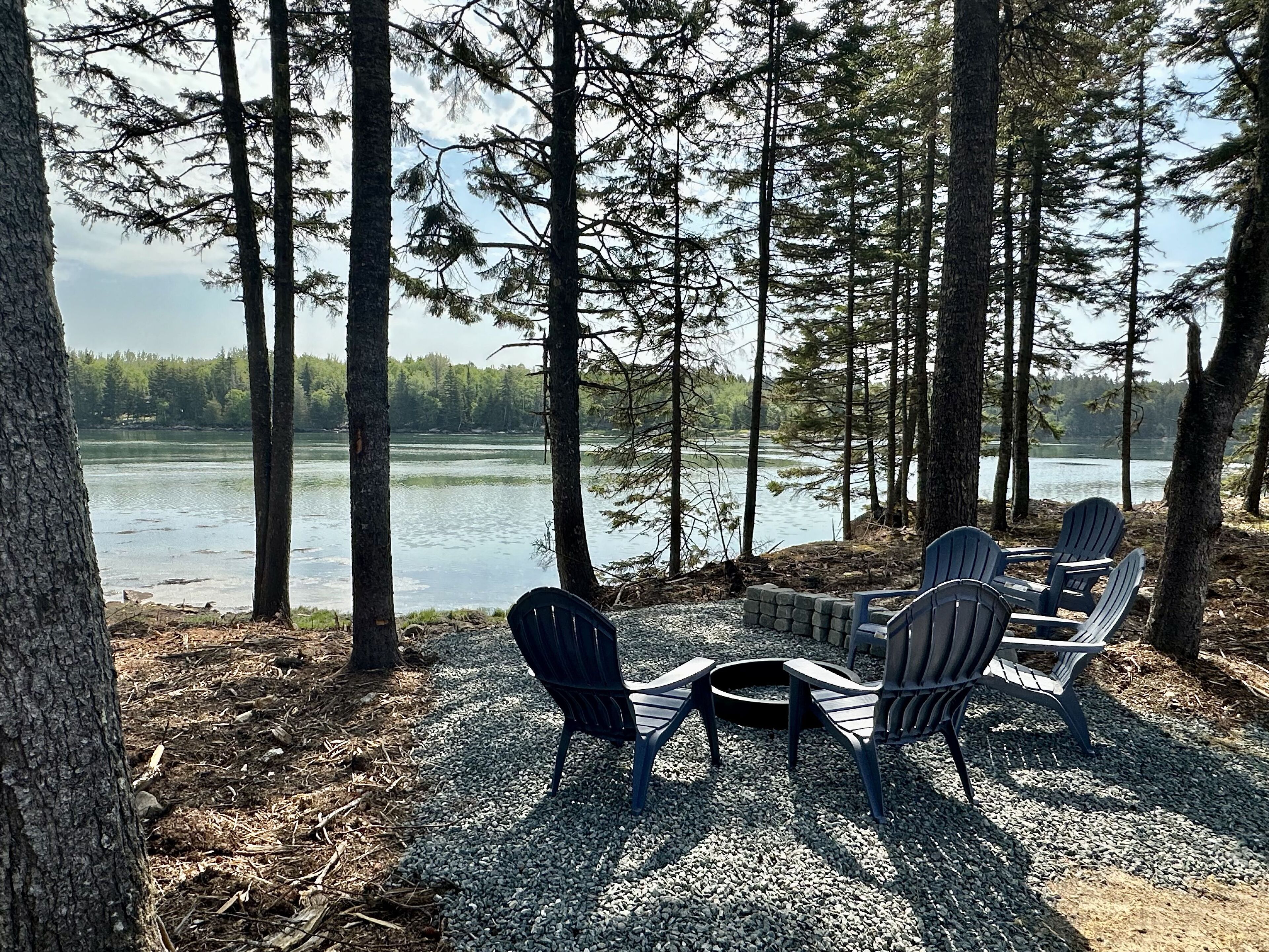Un espacio de descanso junto al lago con sillas Adirondack y una fogata, rodeado de altos árboles y vistas a un lago tranquilo.
