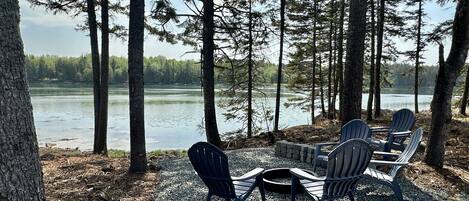 Un espacio de descanso junto al lago con sillas Adirondack y una fogata, rodeado de altos árboles y vistas a un lago tranquilo.