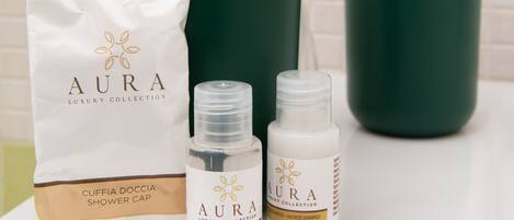 Un baño con pared de azulejos blancos, un lavamanos verde y una bolsa de gorro de ducha blanca con la etiqueta "AURA LUXURY COLLECTION", junto a dos botellas de champú de ducha "AURA LUXURY COLLECTION".
