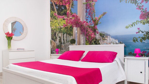 A bedroom with a white bed, pink pillows, and a view of a coastal town. - De Vivo Realty - Stanza Tasso Sorrento (Sorrento)