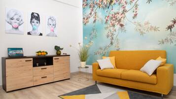 Ein Wohnzimmer mit einem gelben Sofa, einem Holzkasten und einer floralen Wandmalerei.