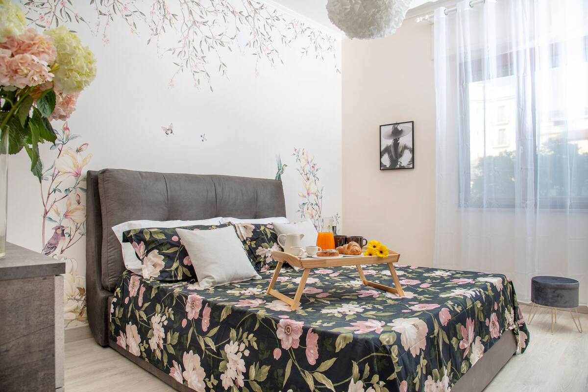A bedroom with a floral bedspread, a grey headboard, a bedside table with a vase of flowers, and a breakfast tray with orange juice and croissants.