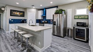A modern kitchen with a central island, bar stools, stainless steel appliances, and a blue backsplash.