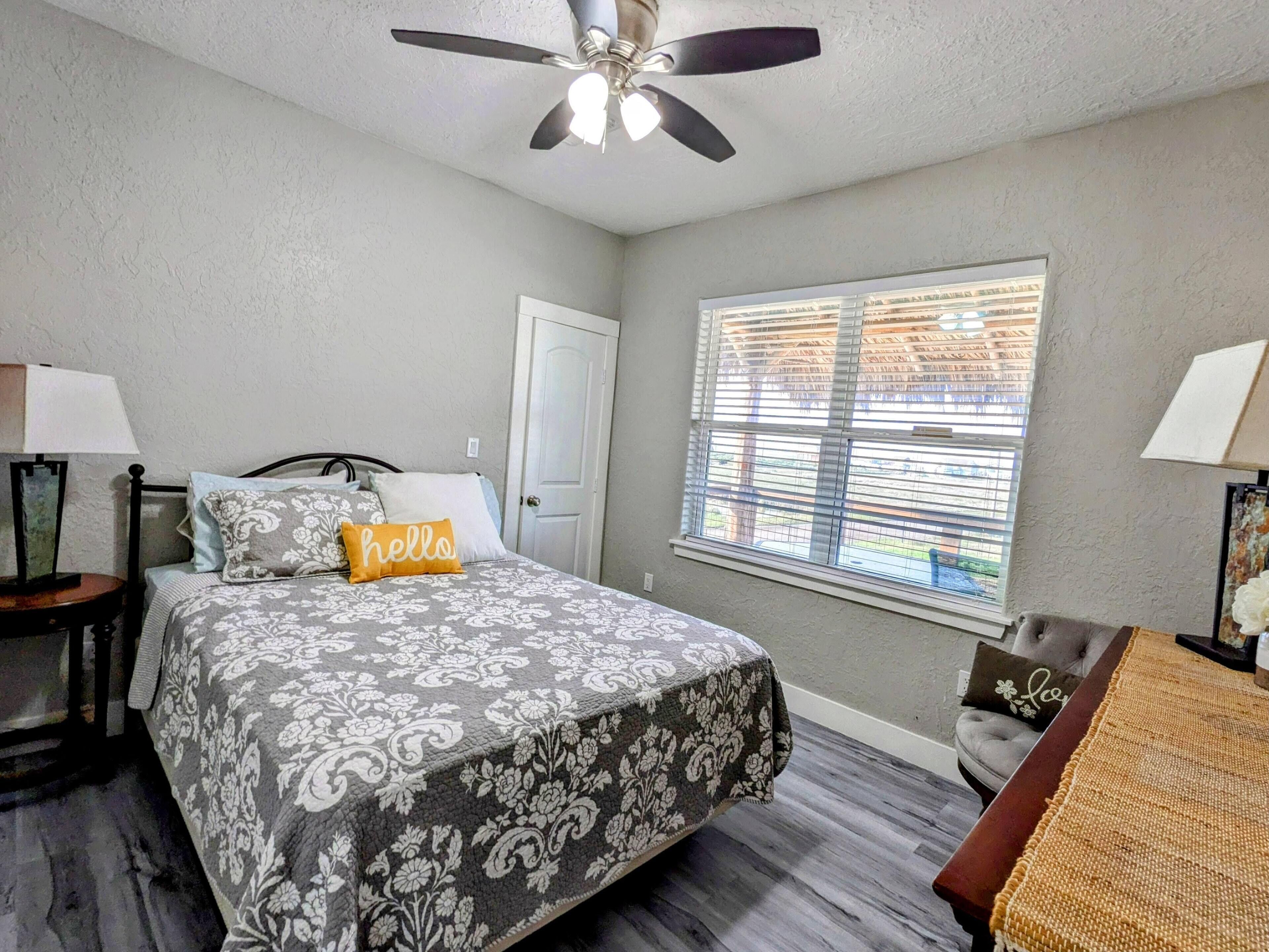A bedroom with a bed, a ceiling fan, a window with blinds, a door, and a nightstand with a lamp.