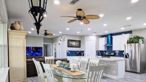 A modern kitchen with a dining area, featuring a glass-top table, white chairs, a ceiling fan, and built-in appliances.