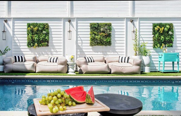 A poolside area with a seating arrangement of two beige sofas with black and white striped cushions, a small round table with a cutting board and watermelon, and a teal chair. - Printer's Mark (Atlanta)