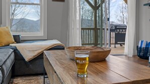 Ett träbord med ett glas öl, en trävasskål och en soffa i ett rum med utsikt över bergen, och en grill utomhus.