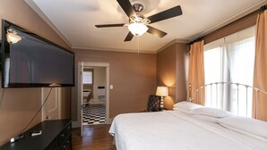 A hotel room with a bed, a ceiling fan, a TV mounted on the wall, a desk with drawers, and a window with curtains. - Upper 2 Bedroom Loft Apartment (Memphis)