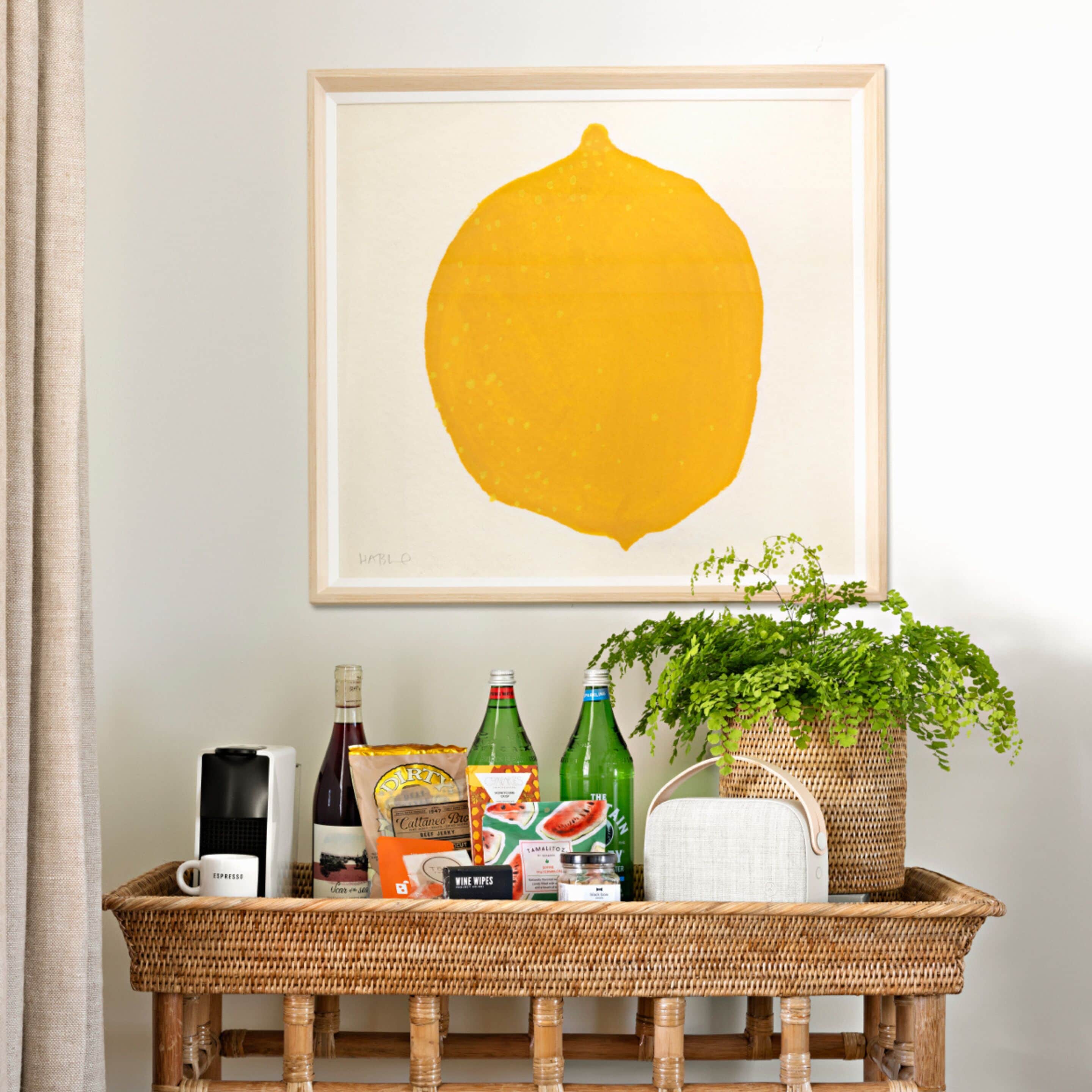 A wicker table with a framed artwork, a bottle of wine, a bag of snacks, a bottle of water, a plant, and a coffee maker.