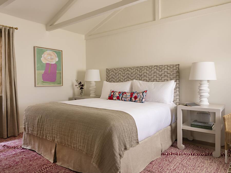 Un dormitorio ordenado con una cama grande, dos mesitas de noche con lámparas, una silla y una obra de arte en la pared.