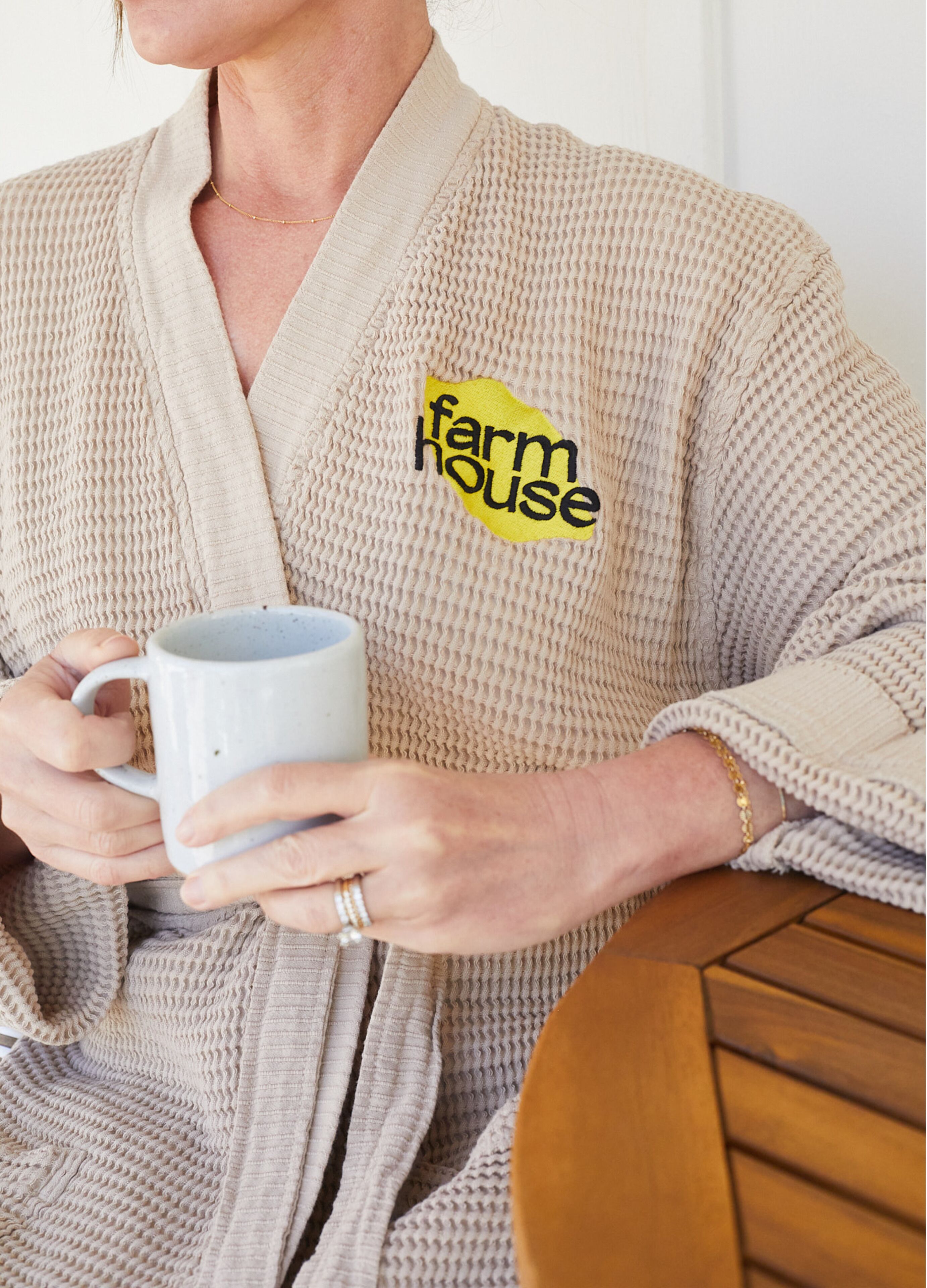 Una persona in una veste beige tiene una tazza bianca, con il logo 'farmhouse' sulla veste.