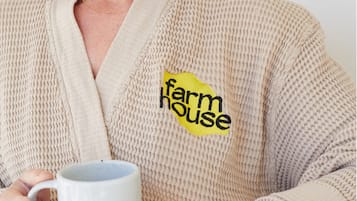 Una persona in una veste beige tiene una tazza bianca, con il logo 'farmhouse' sulla veste.