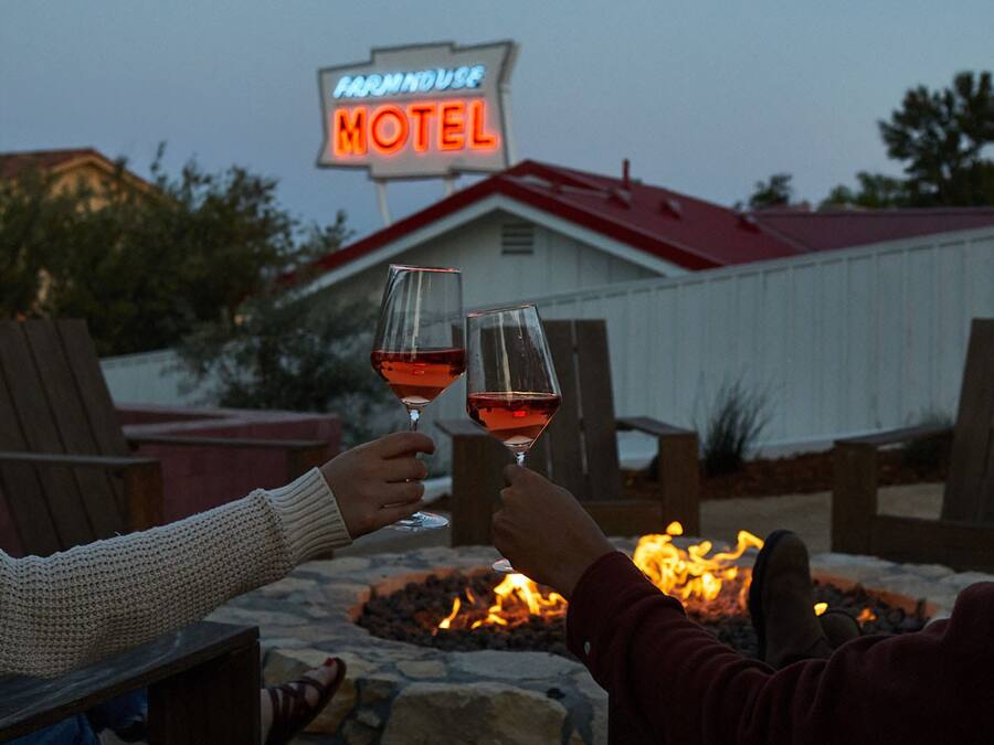 Two people toasting with wine glasses at a fire pit with a 'Farmhouse Motel' sign in the background.