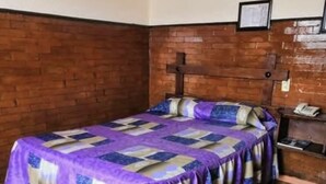 A single bed with a purple and beige quilt, a wooden headboard, and a brick wall. - Hotel San Agustin (Puebla)