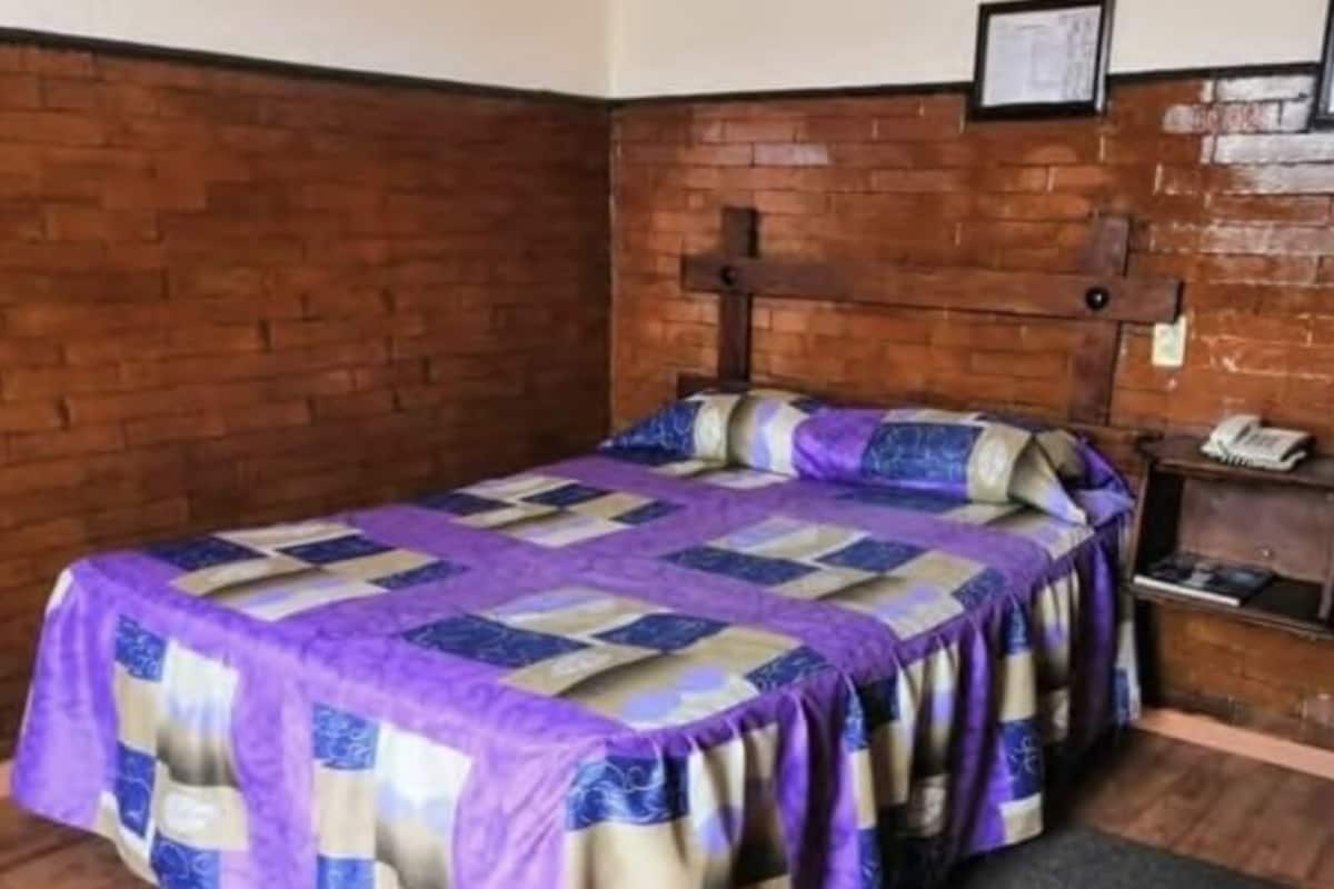 A single bed with a purple and beige quilt, a wooden headboard, and a brick wall.