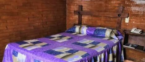 A single bed with a purple and beige quilt, a wooden headboard, and a brick wall.