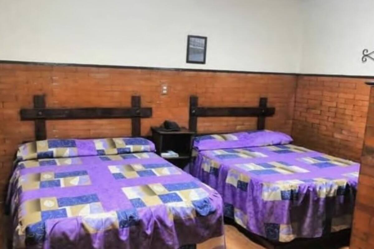 A room with two beds, each with a purple and blue quilt. There is a small table between the beds.