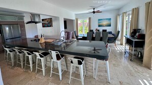 A modern kitchen with a central island, bar stools, a dining area with chairs, a refrigerator, and a ceiling fan.